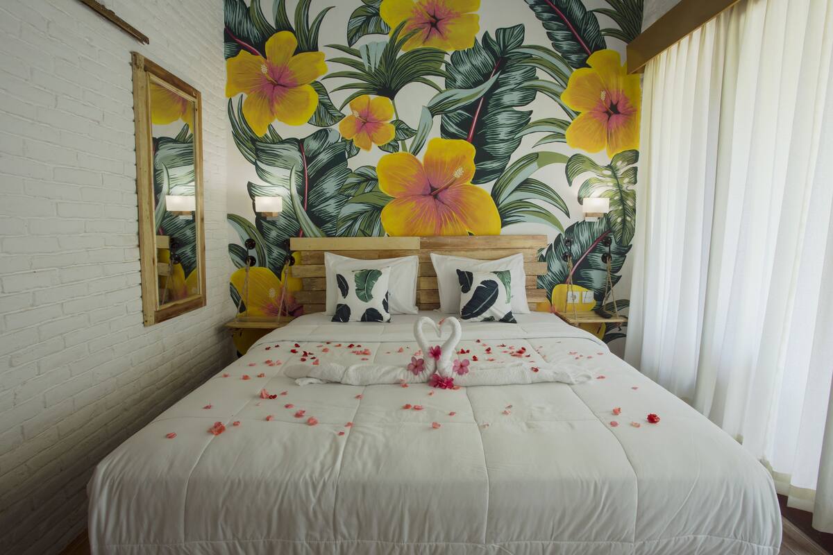 Une chambre d’hôtel avec un grand lit, des murs blancs, une fresque murale à motifs floraux et une tête de lit en bois.