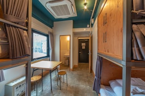 A compact room with bunk beds, a small table, and a chair. There is a window with curtains, a door leading to another room, and an air conditioning unit on the ceiling.