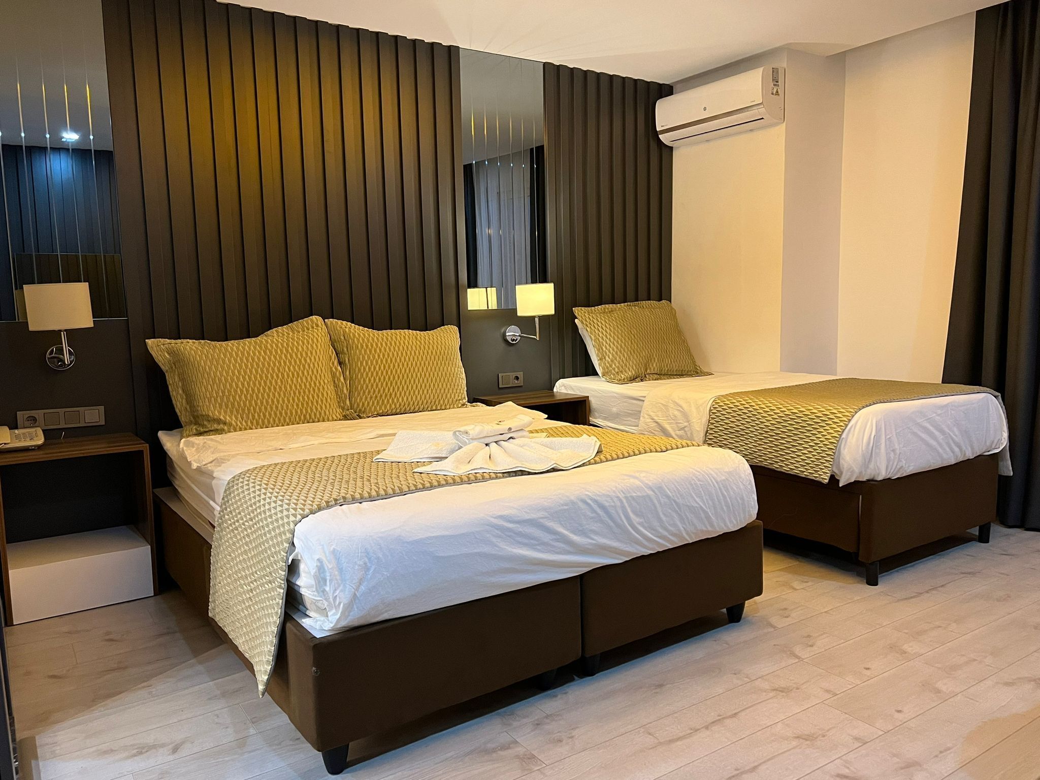 A hotel room with two beds, a bedside table with a lamp, a wall-mounted air conditioner, and a mirror.