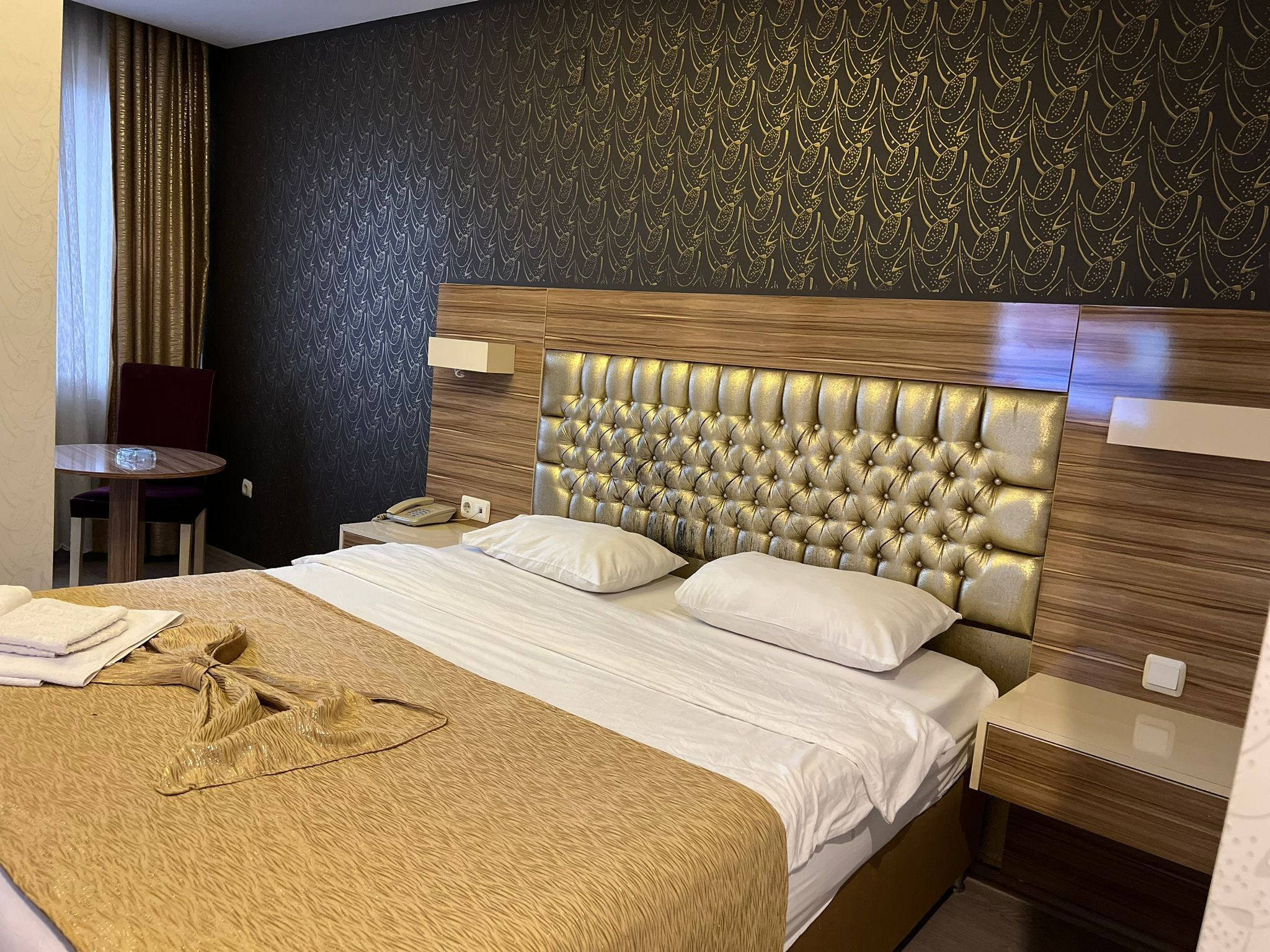 A hotel room with a large bed, a bedside table, a chair, and a patterned wall.