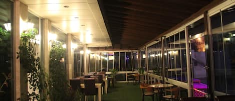 An indoor dining area with tables and chairs, large windows, and potted plants.