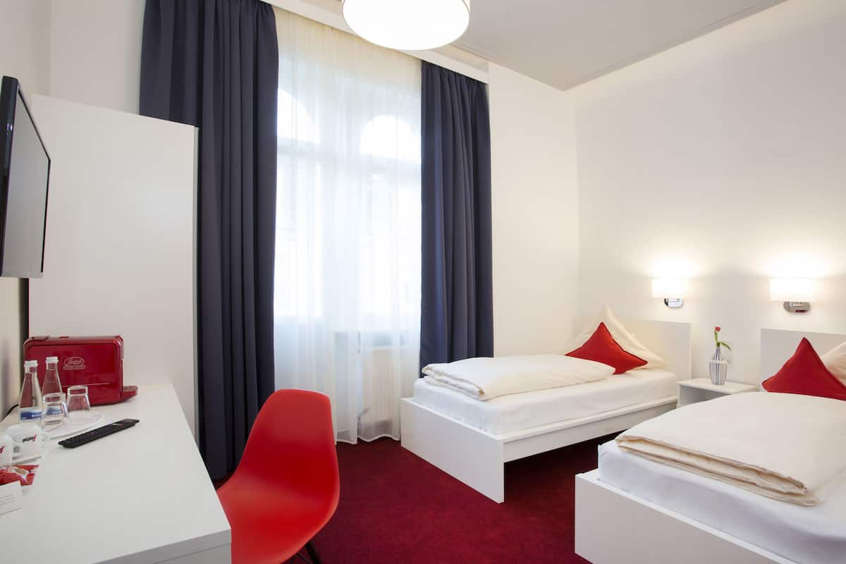 Une chambre d’hôtel avec un bureau blanc, une chaise rouge, deux lits avec des oreillers rouges, une valise rouge et un vase avec une fleur.