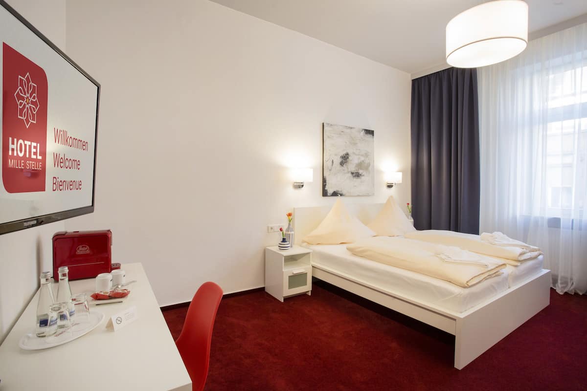 Une chambre d’hôtel avec un lit, des tables de chevet, un bureau et une chaise. On peut voir une valise rouge sur le bureau et un panneau d’accueil encadré accroché au mur.