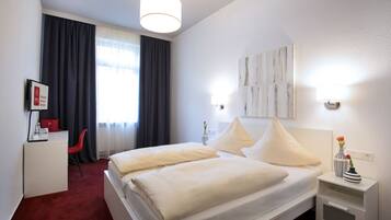A hotel room with a bed, a desk, a chair, a nightstand, and a window with curtains.