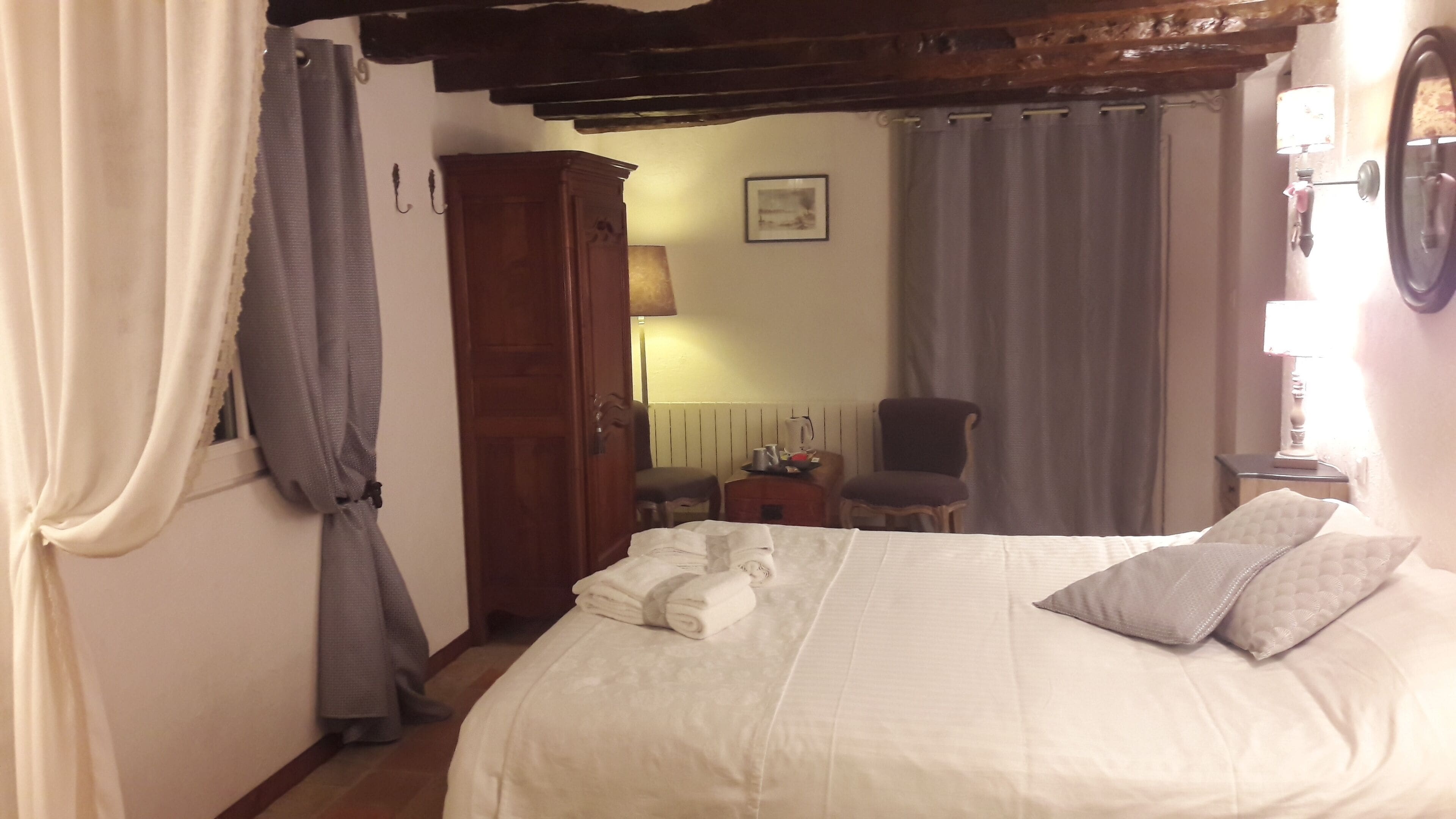 Uma cama bem arrumada com almofadas, um guarda-roupa de madeira, uma cadeira e uma mesa com abajur em um quarto com cortinas e um quadro na parede.