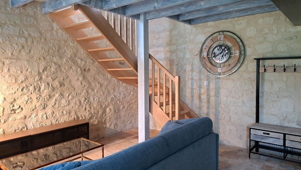 Un interior rústico con pared de piedra, escalera de madera, reloj metálico redondo, sofá azul y una mesita.