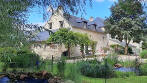 Une maison en pierre, dotée d’un toit à forte pente, entourée d’une végétation luxuriante et d’un petit étang.