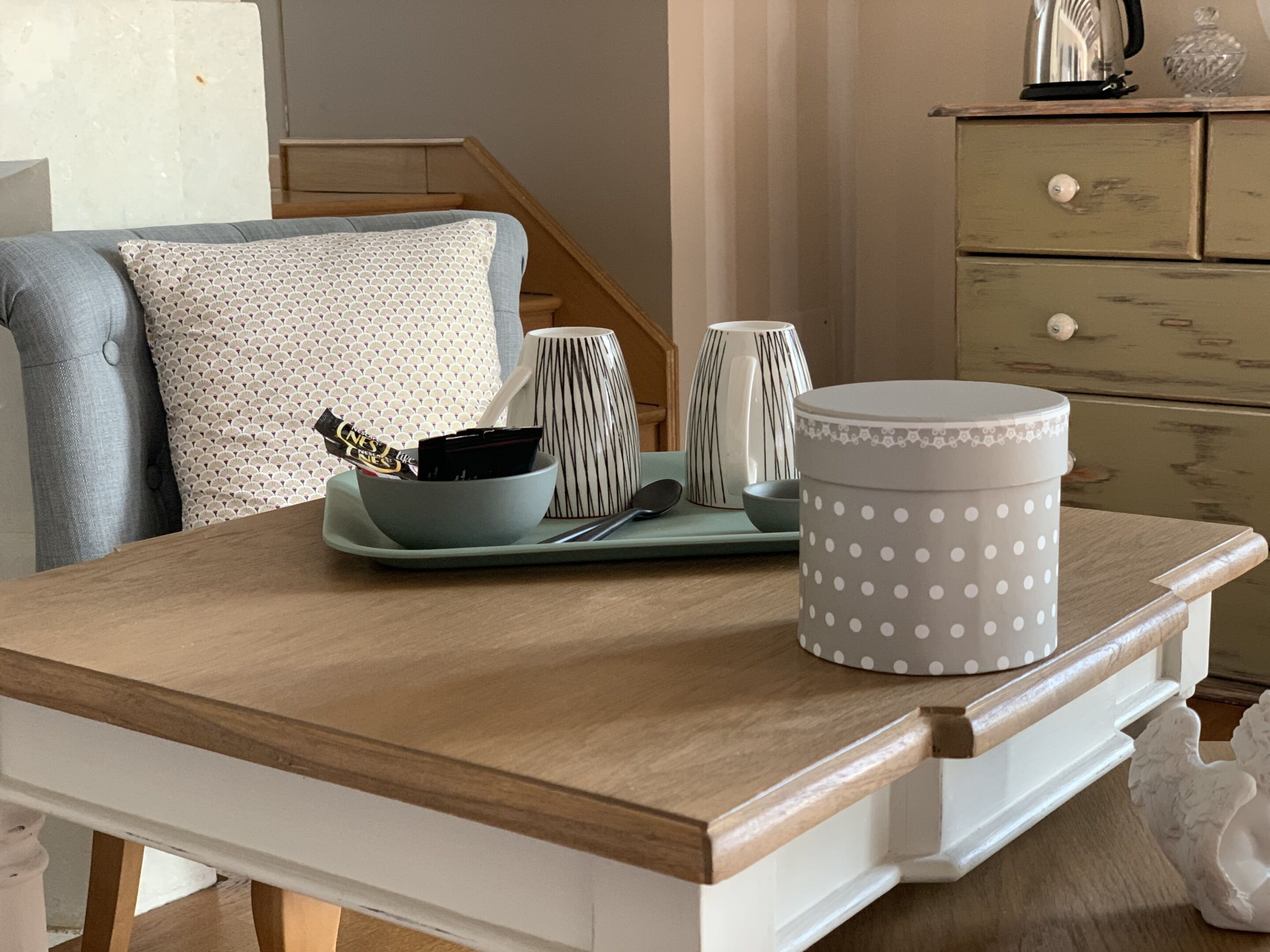 Una mesa de madera con un recipiente gris de lunares, una taza a rayas y un bol con una barra de chocolate.