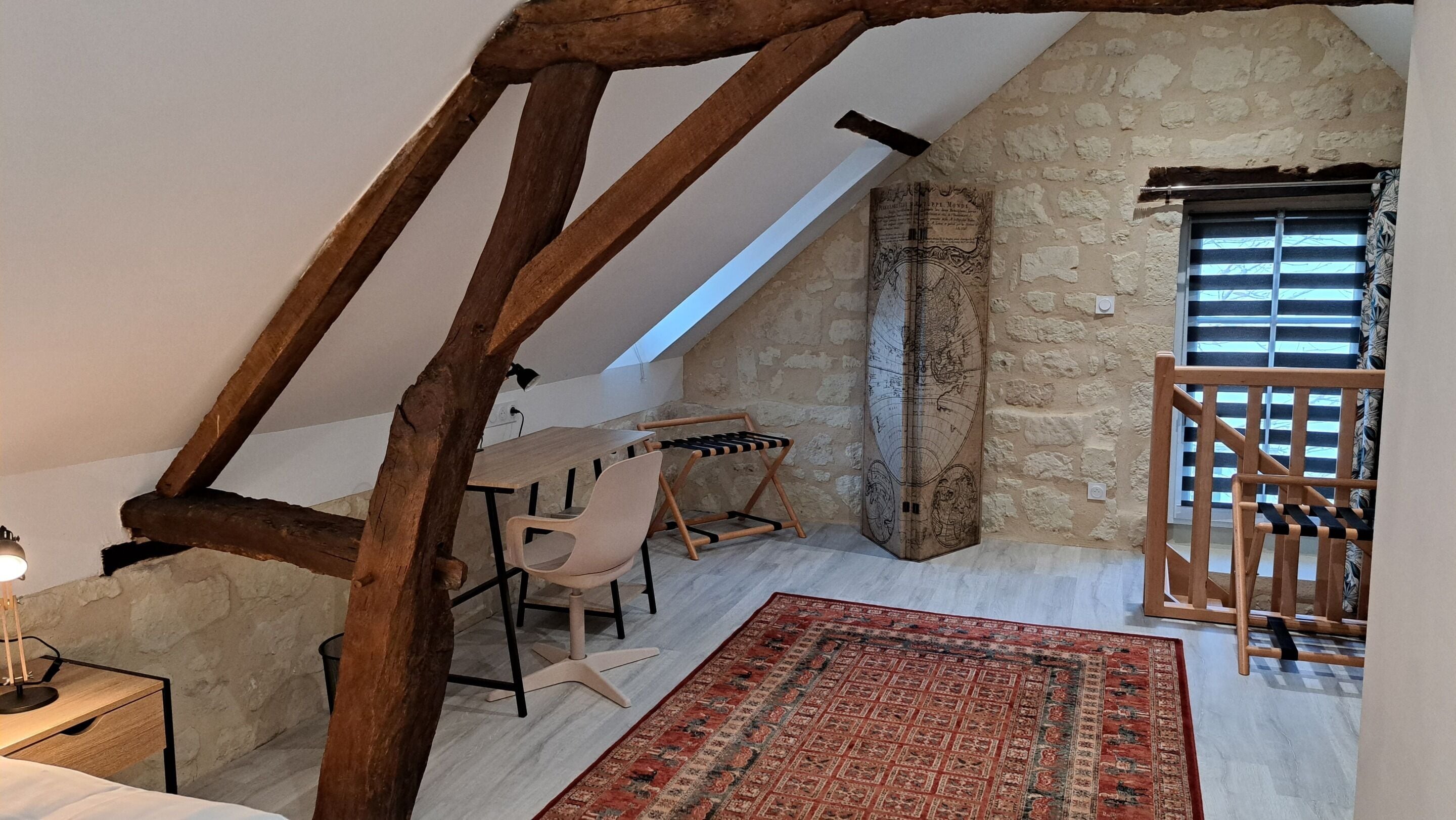 A loft bedroom with exposed wooden beams, a stone wall, a large map, a wooden chair, and a red patterned rug.
