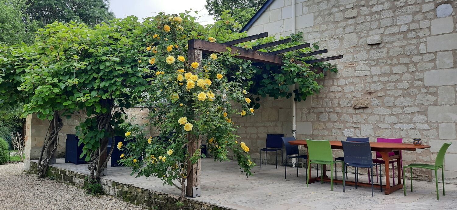 A stone building with a pergola covered in yellow flowers, outdoor seating with colorful chairs, and a gravel path.
