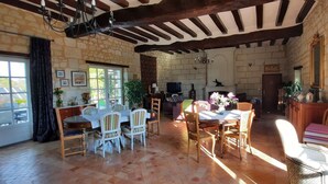 Un vaste salon doté d’un mur en pierre, de poutres apparentes, d’une cheminée, d’un coin repas avec des chaises et d’un téléviseur.