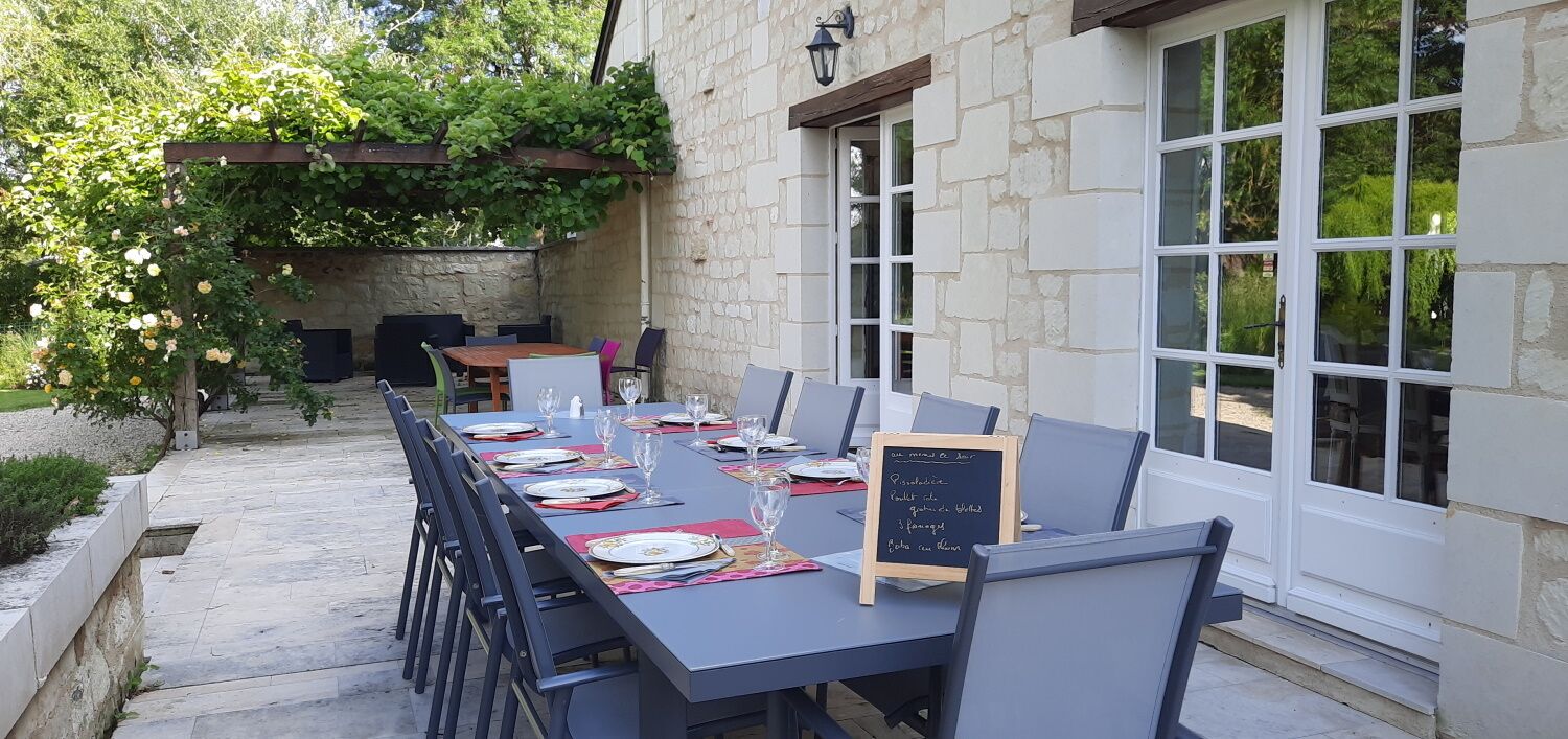 Zona pranzo all'aperto con parete in pietra, ampie finestre e una pergola con piante rampicanti.