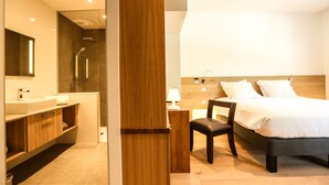 A modern hotel room with a wooden headboard, a large bed, a bedside table, a chair, a desk, a mirror, a bathroom with a sink and shower. - Le Kube Annecy Appartements De Luxe (Annecy)