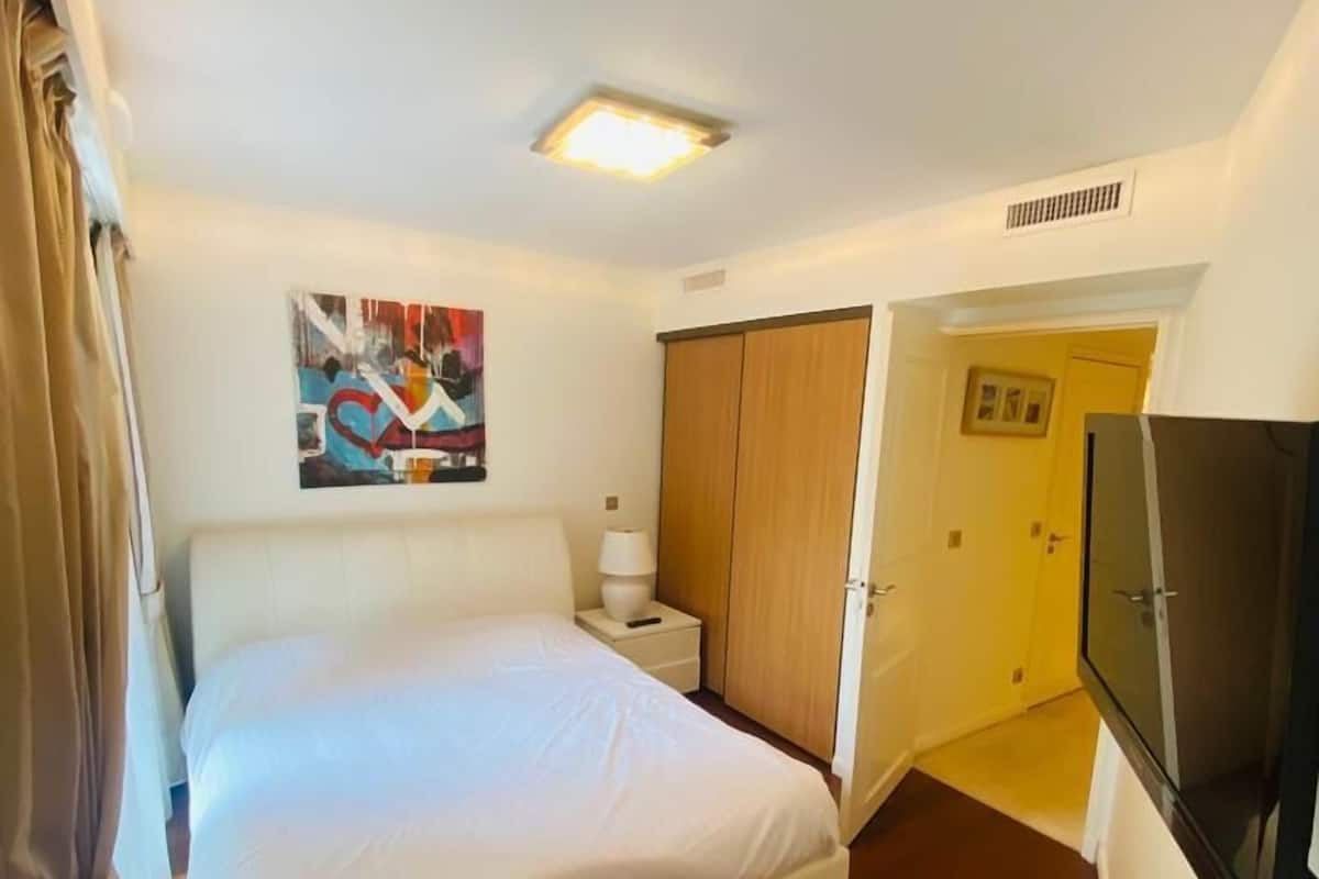 Habitación de hotel con cama, mesita de noche, lámpara, un cuadro, armario, televisor y una puerta que da a otra habitación.