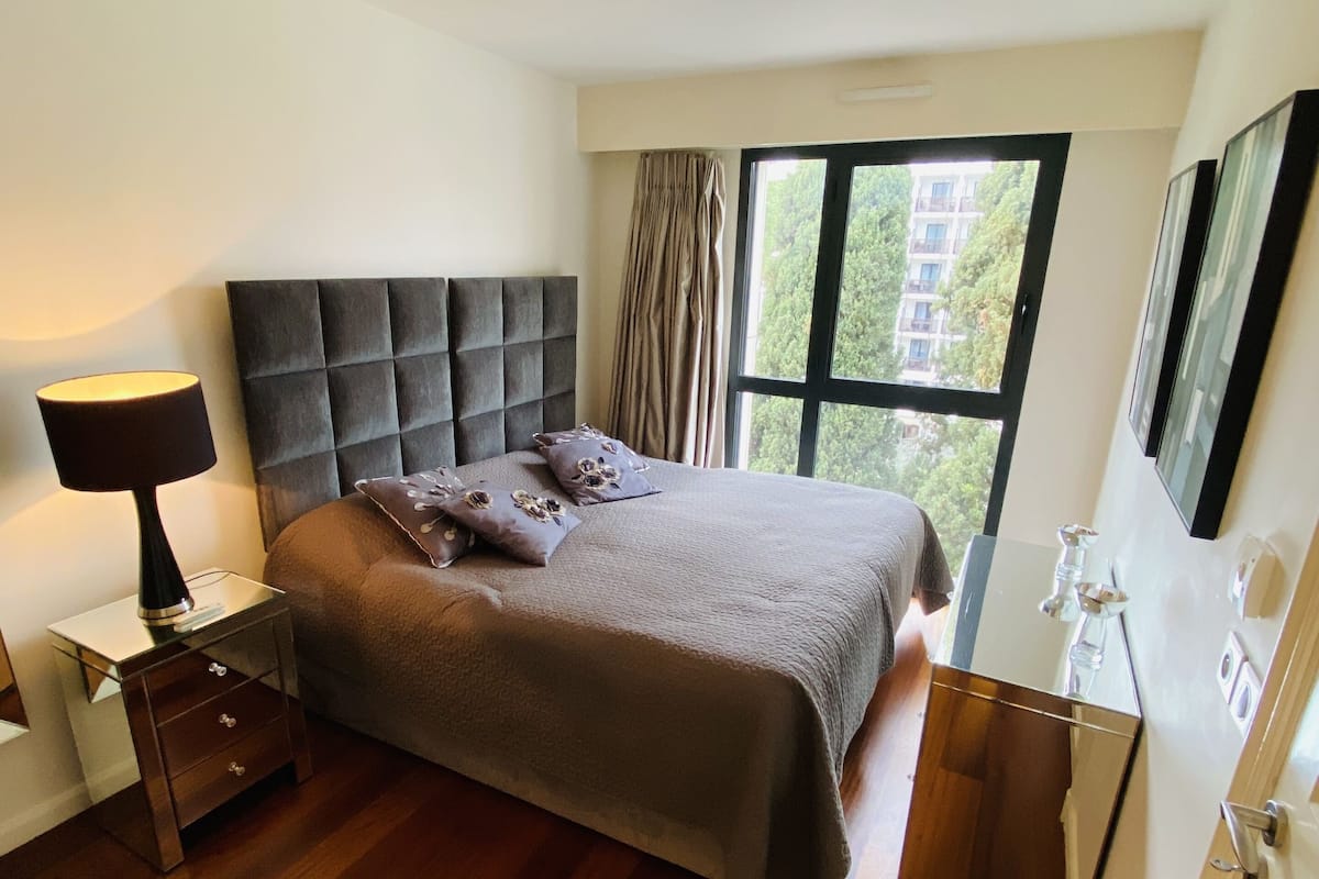 Un dormitorio con una cama grande, una mesita de noche con lámpara, una ventana con cortinas y un espejo en la pared.