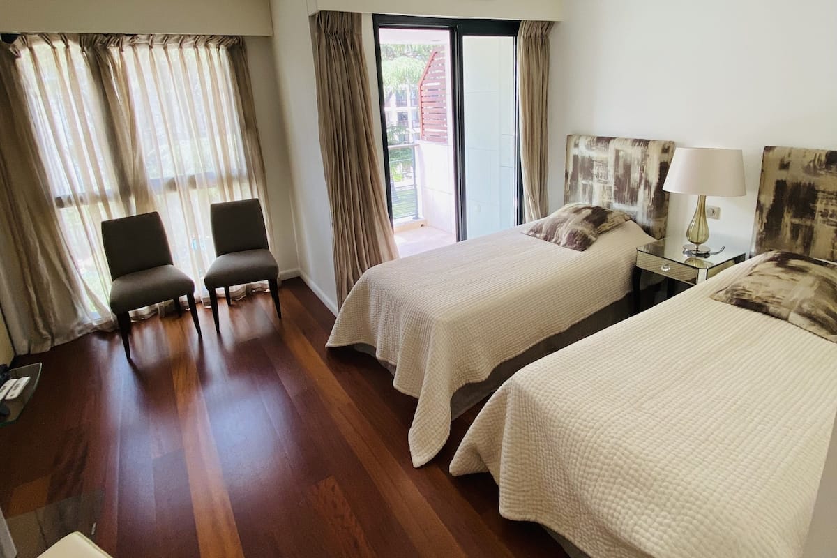 Habitación de hotel con dos camas, un sofá, una silla, una mesita de noche con una lámpara y vistas al exterior a través de una puerta corrediza.