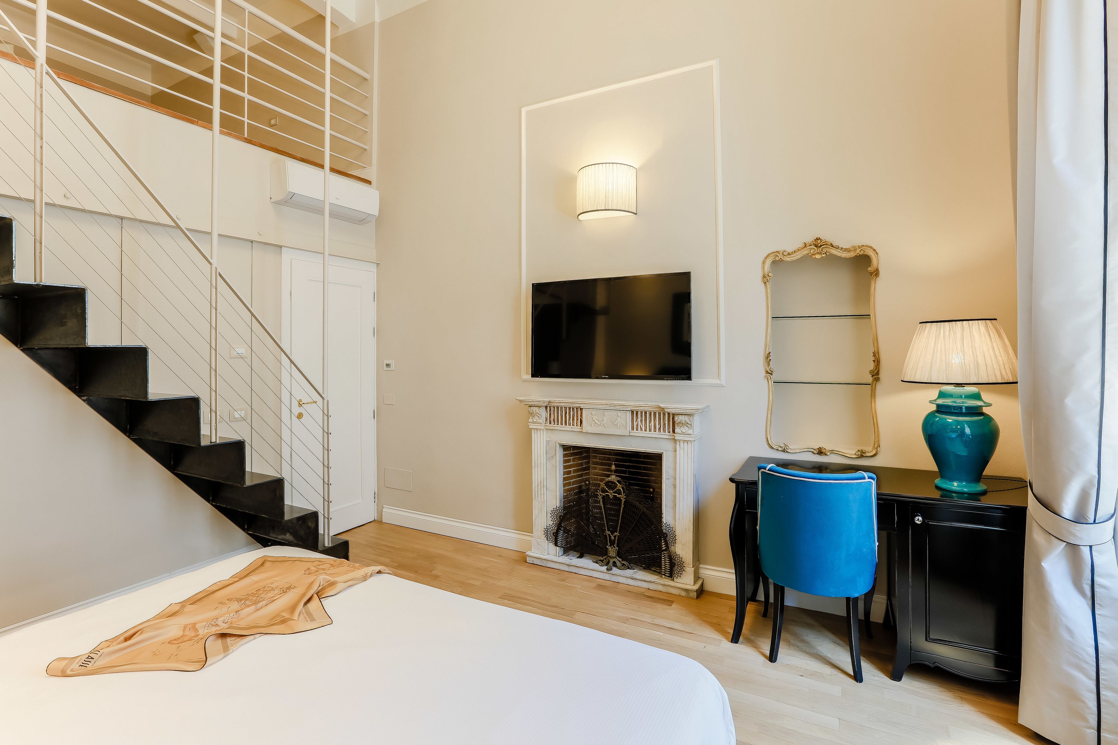 Une chambre à coucher comprenant un lit, un téléviseur fixé au-dessus d’une cheminée, une chaise et un bureau avec une lampe.
