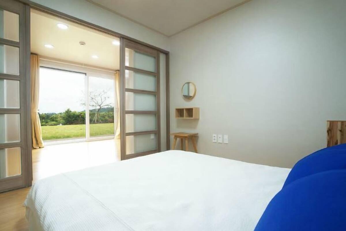 A bedroom with a large bed, a wooden headboard, a small wooden stool, a round mirror on the wall, and a view of a green landscape through a sliding glass door.