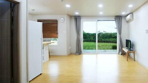 Una habitación amplia con suelo de madera, un ventanal corredero que da a un jardín, una mesita con una silla y un televisor sobre un mueble.