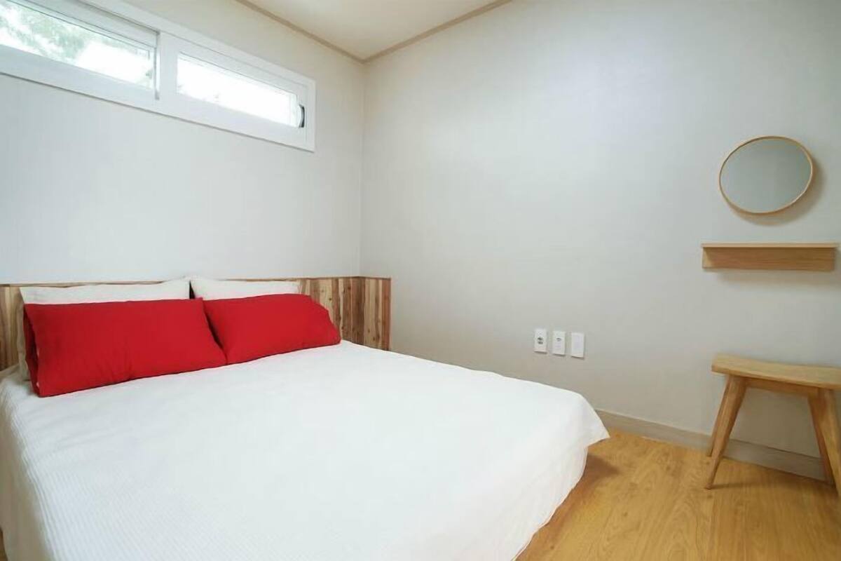 A bedroom with a bed, red pillows, a wooden headboard, a small wooden stool, a round mirror on the wall, and a window with blinds.