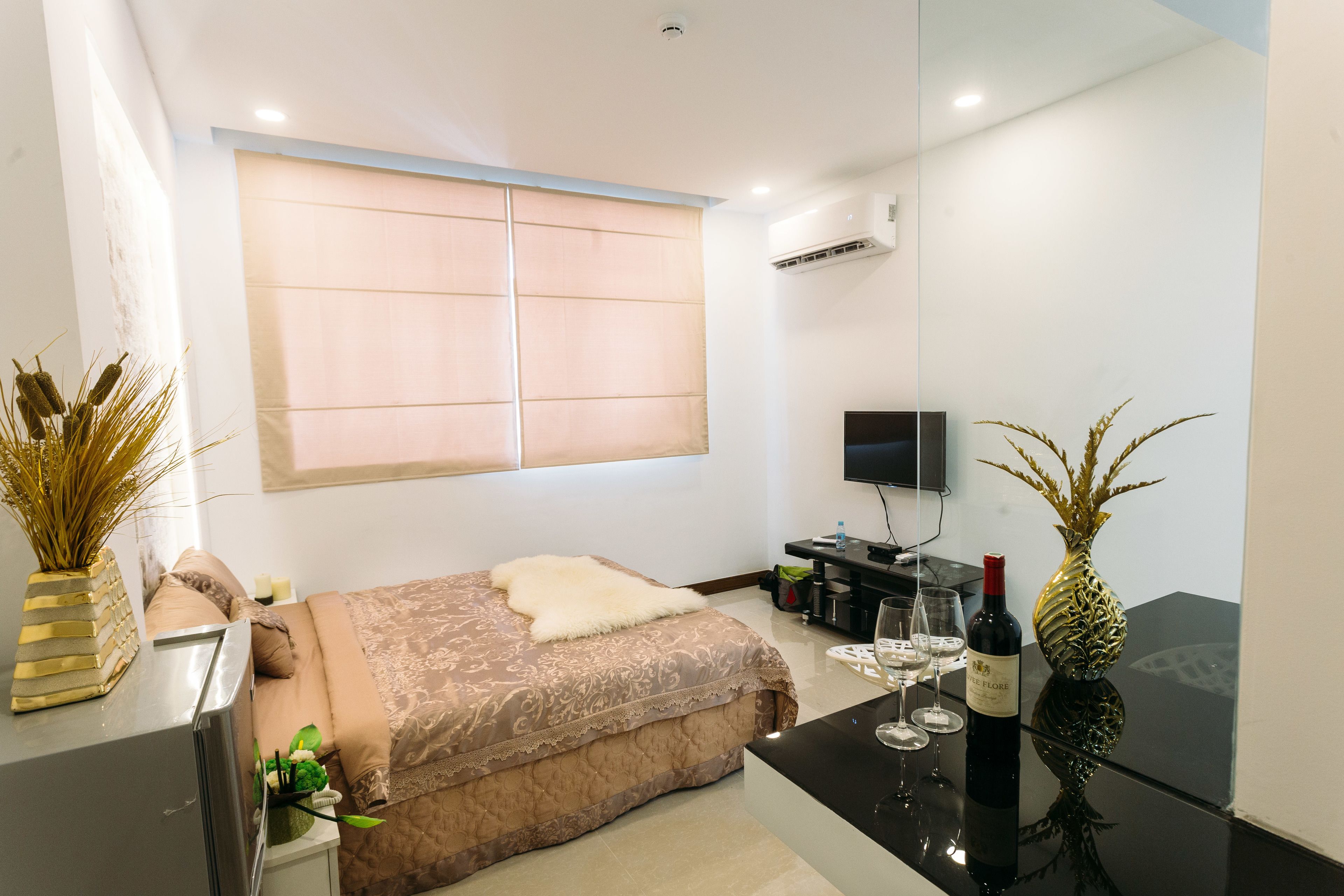 A modern bedroom with a large bed, a flat-screen TV, a small table with a wine bottle and glasses, and decorative plants.