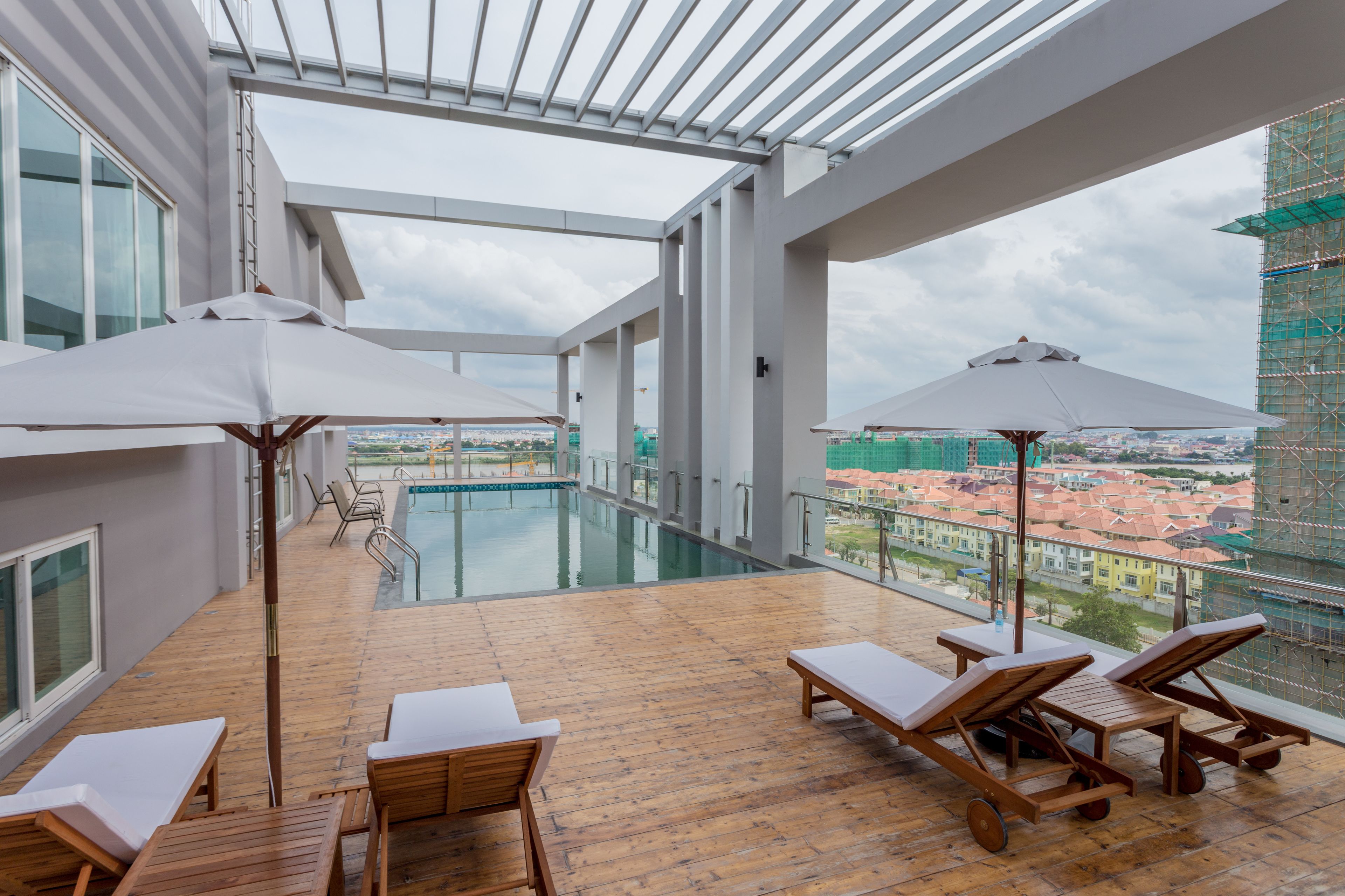 A rooftop pool area with wooden lounge chairs and umbrellas, a glass-enclosed swimming pool, and a view of the cityscape.