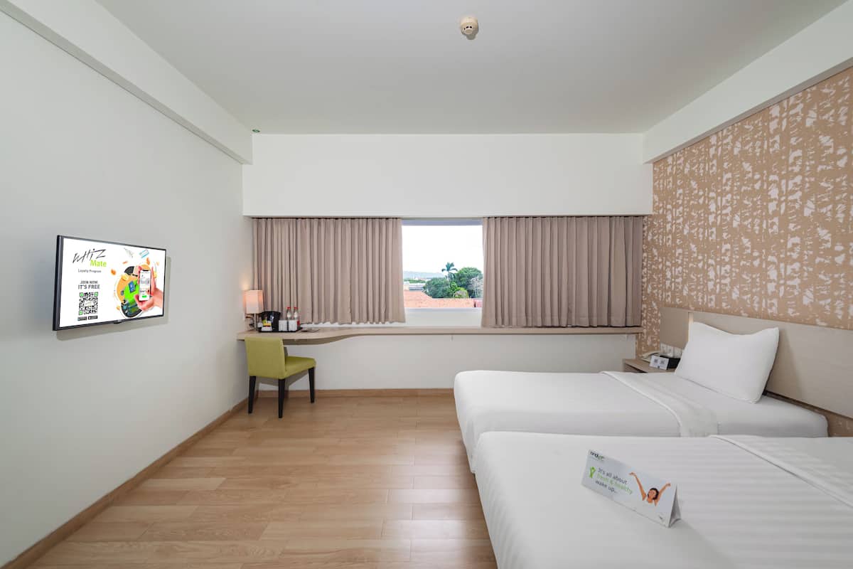 Ein Hotelzimmer mit zwei Betten, einem Schreibtisch mit Stuhl, einem Fenster mit Vorhängen und einem an der Wand montierten Fernseher, der eine Werbung zeigt.