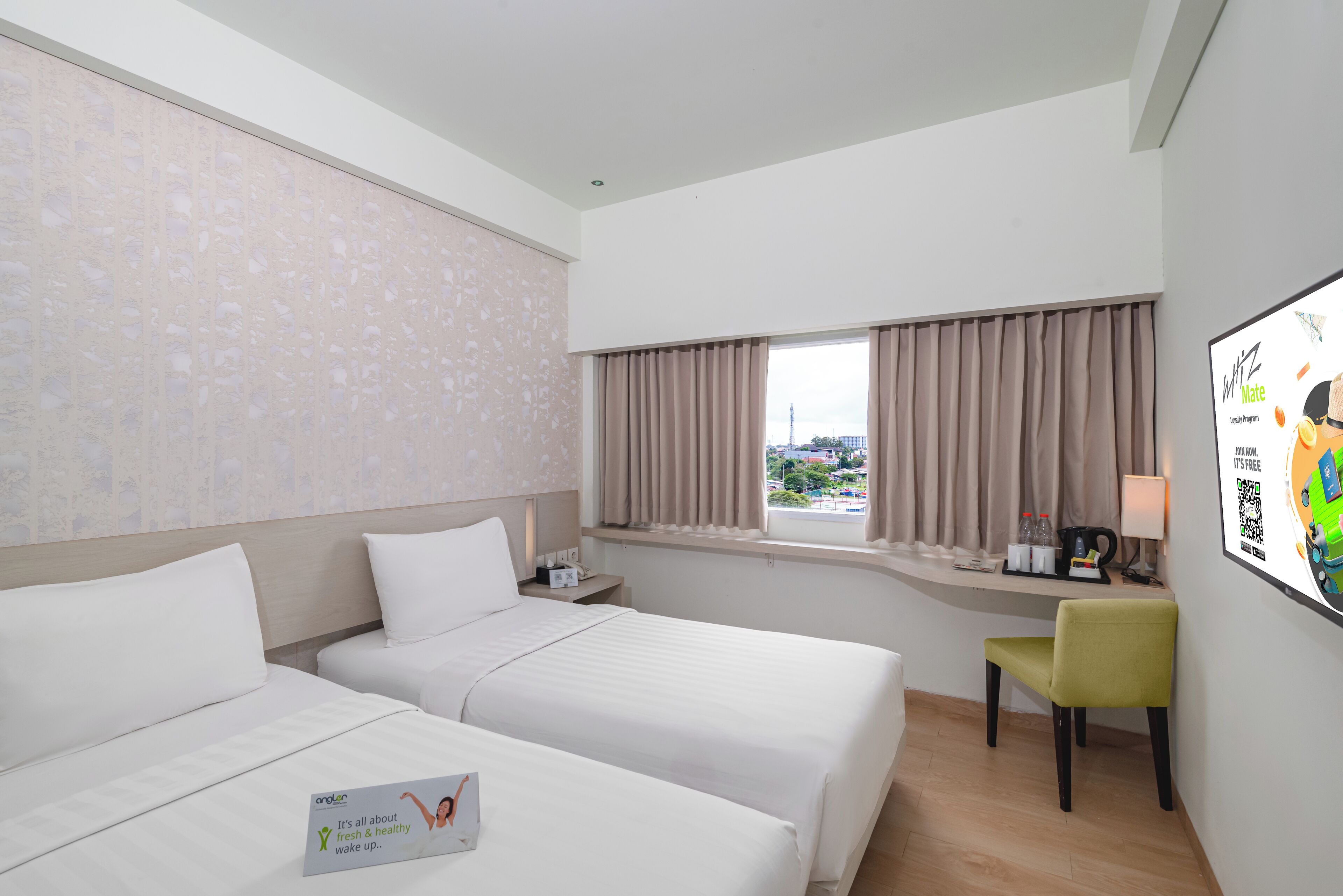 A hotel room with two beds, a desk, a chair, a window with curtains, and a wall-mounted screen displaying a loyalty program.
