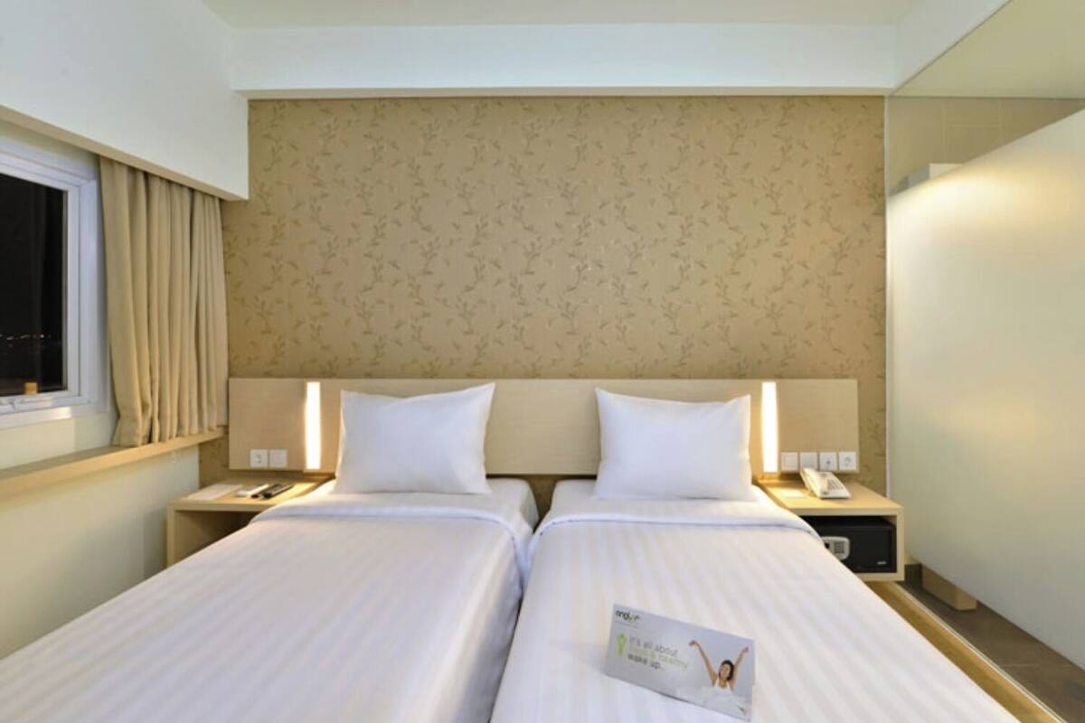 Ein Hotelzimmer mit zwei Betten, einem Fenster mit Vorhängen, einem Kopfteil mit Blumenmuster, Nachttischen mit Lampen und einer Zeitschrift auf dem Bett.