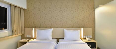A hotel room with two beds, a window with curtains, a headboard with a floral pattern, bedside tables with lamps, and a magazine on the bed.