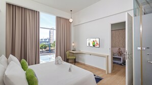 Une chambre d’hôtel comprenant un lit, un bureau, une chaise, une fenêtre avec des rideaux, un téléviseur fixé au mur et une porte donnant sur une autre pièce.