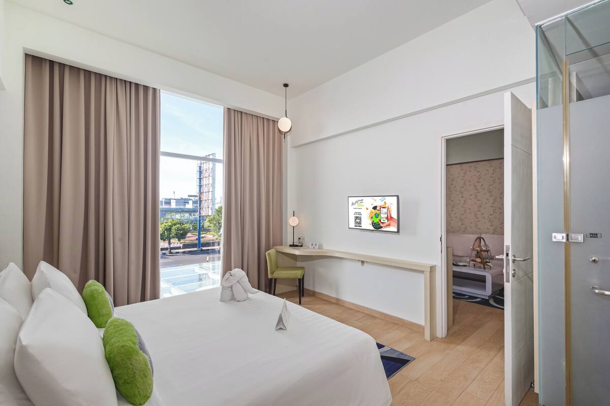 Ein Hotelzimmer mit Bett, Schreibtisch, Stuhl, einem Fenster mit Vorhängen, einem an der Wand befestigten Fernseher und einer Tür zu einem weiteren Raum.