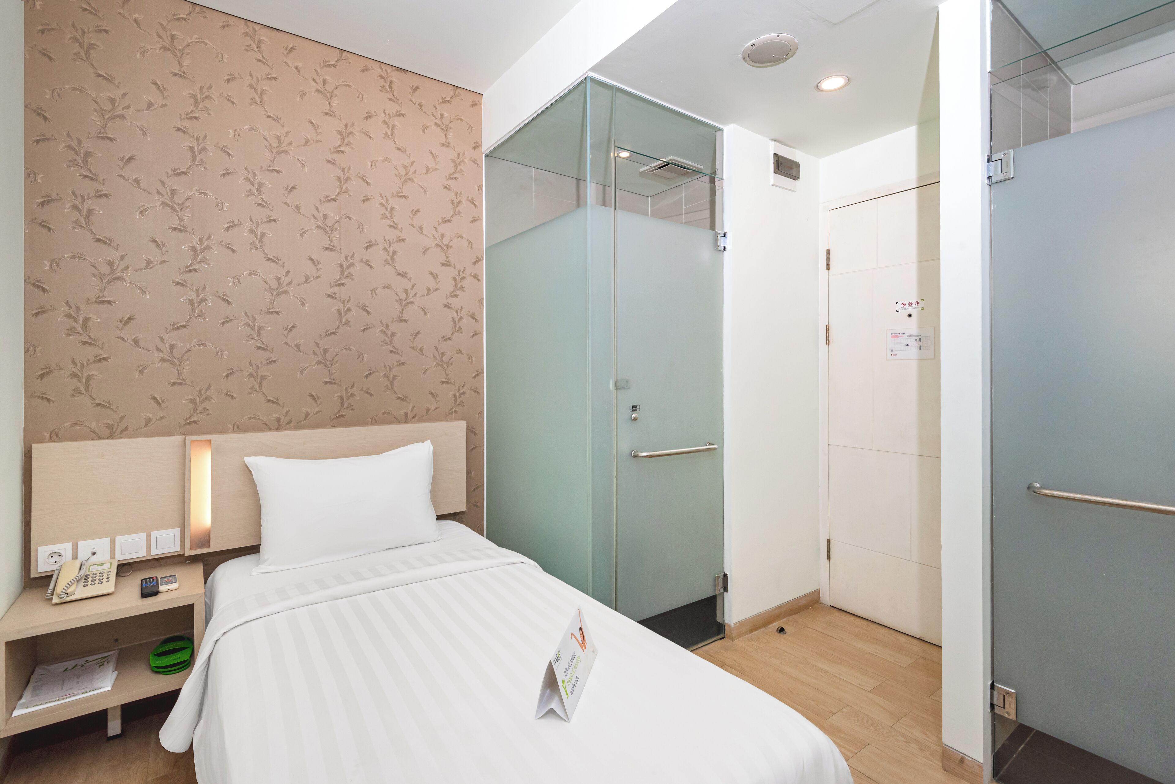 A hotel room with a bed, a nightstand with a phone and a remote, a glass-enclosed shower, and a wardrobe.