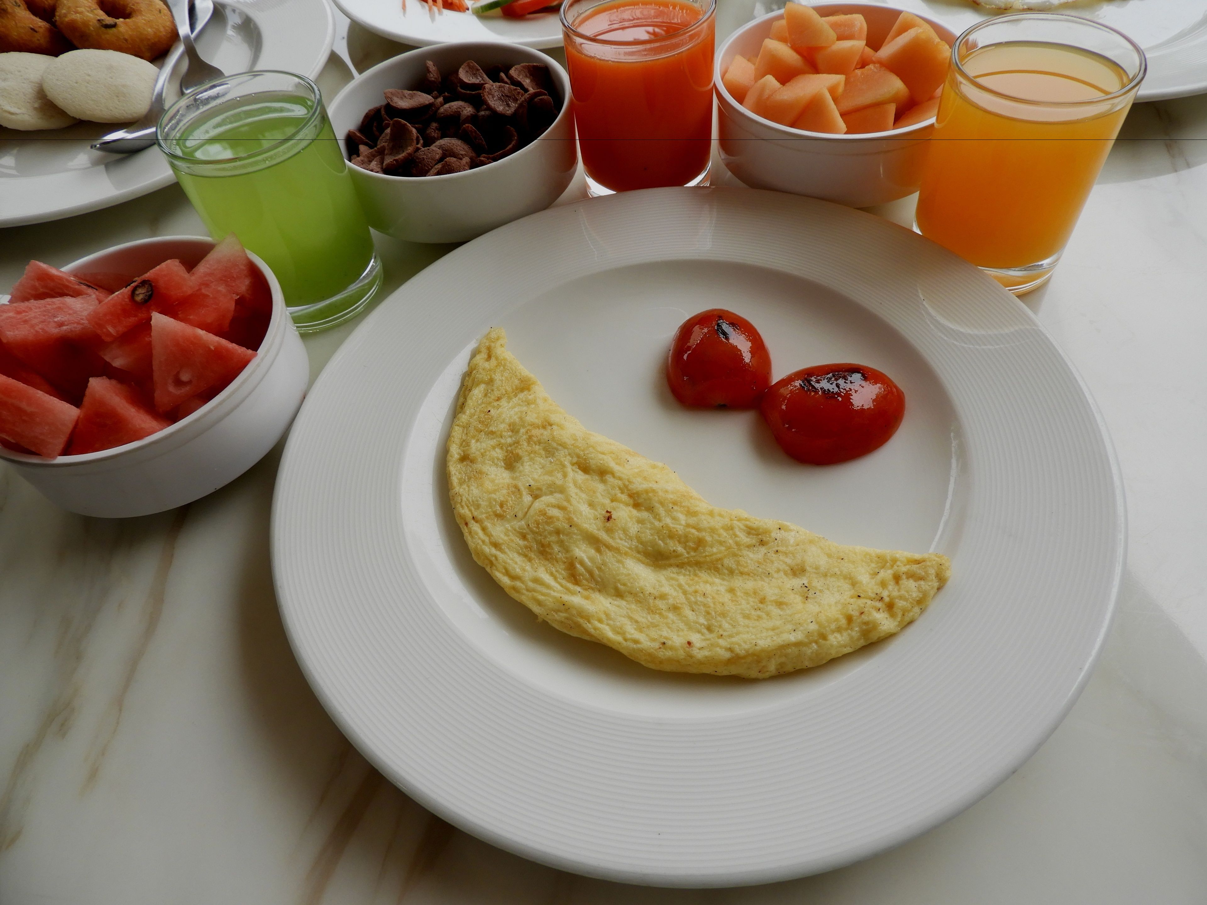 A breakfast plate with an omelette, watermelon, cherry tomatoes, and various beverages.