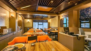Een restaurant met houten tafels en oranje stoelen, een balie met een computer, en uitzicht op de straat.