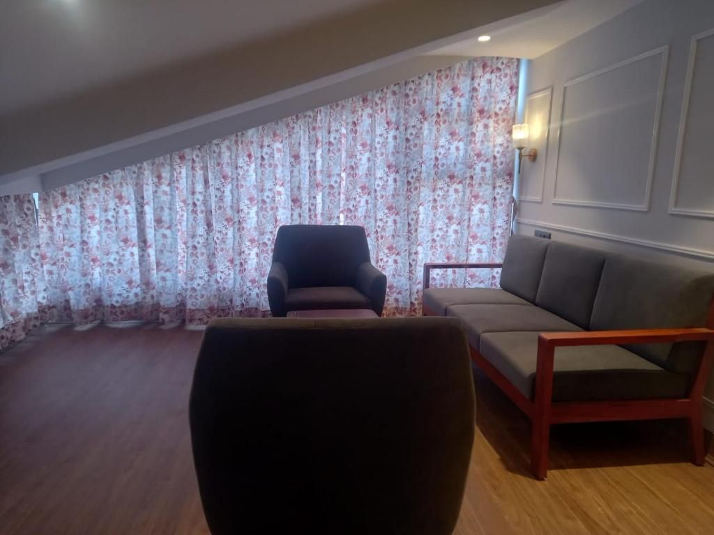 A room with a wooden floor, a dark armchair, a grey sofa, and a wall with a floral curtain.