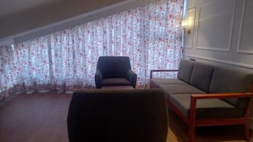 A room with a wooden floor, a dark armchair, a grey sofa, and a wall with a floral curtain.