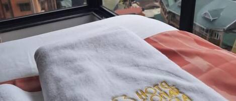 Una cama con una toalla blanca y un edredón rojo, una ventana con vistas a un paisaje urbano.