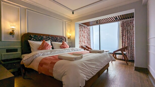 Een hotelkamer met een groot bed, twee bedlampen, een houten hoofdbord, een raam met gordijnen en een kleine tafel met een stoel.