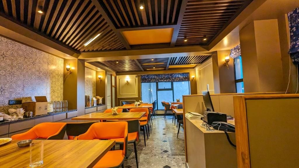 Restaurante com mesas de madeira e cadeiras laranjas, parede estampada e vista pela janela.