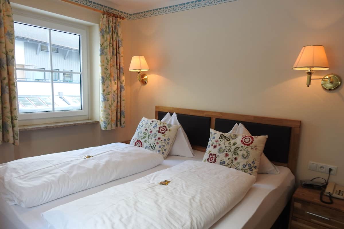 A double bed with floral pillows, two bedside lamps, a window with curtains, and a wooden nightstand with a phone.