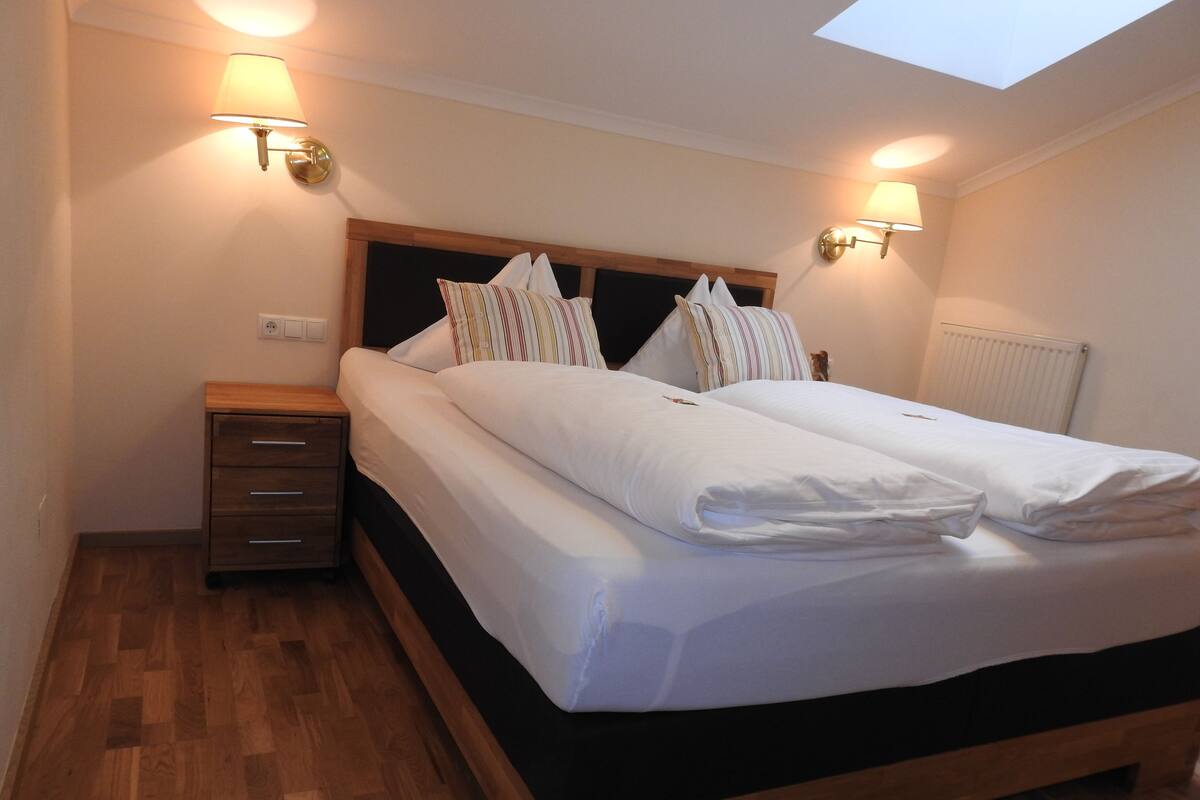 A neatly made bed with white linens and pillows, a wooden headboard, a nightstand with drawers, and two wall-mounted lamps.