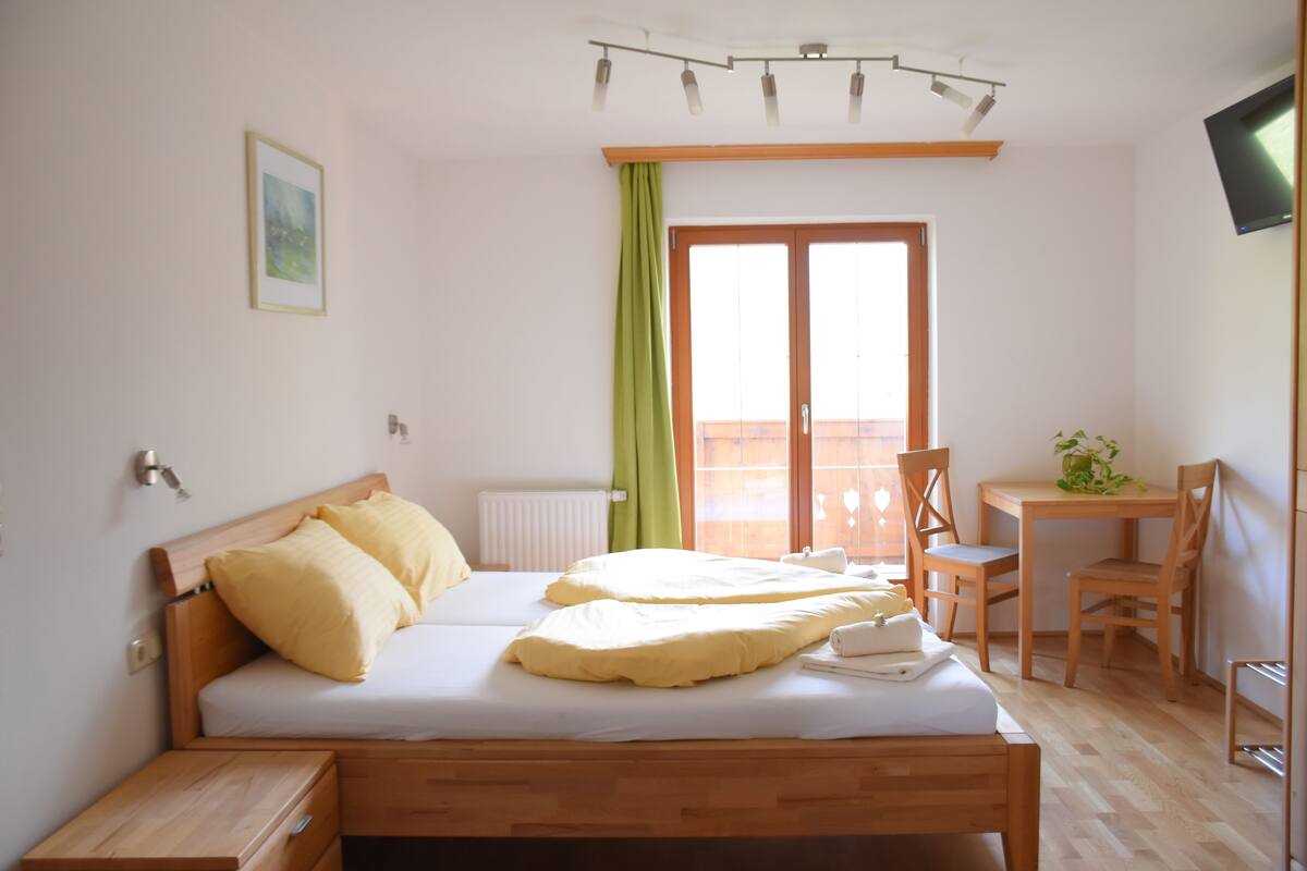 Un dormitorio con cama, mesita de noche, un pequeño escritorio con silla, un radiador y una ventana con cortinas.