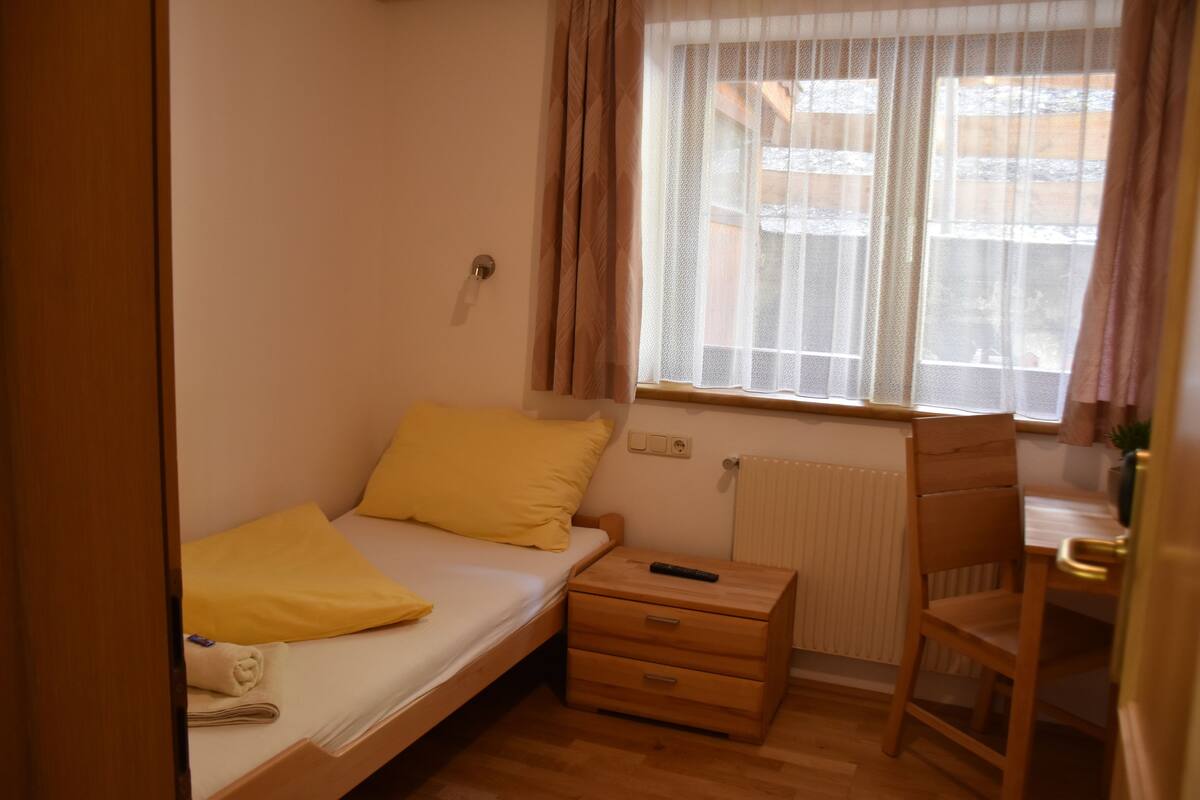 Una habitación pequeña con cama individual, una mesita de noche de madera, una silla y una ventana con cortinas.