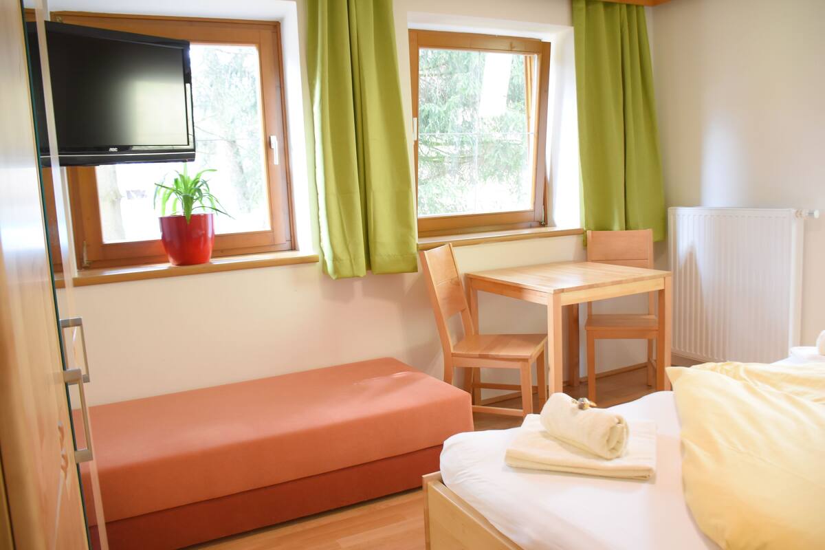 Una habitación con una cama, un escritorio, una silla, un banco, un televisor y una ventana con cortinas.