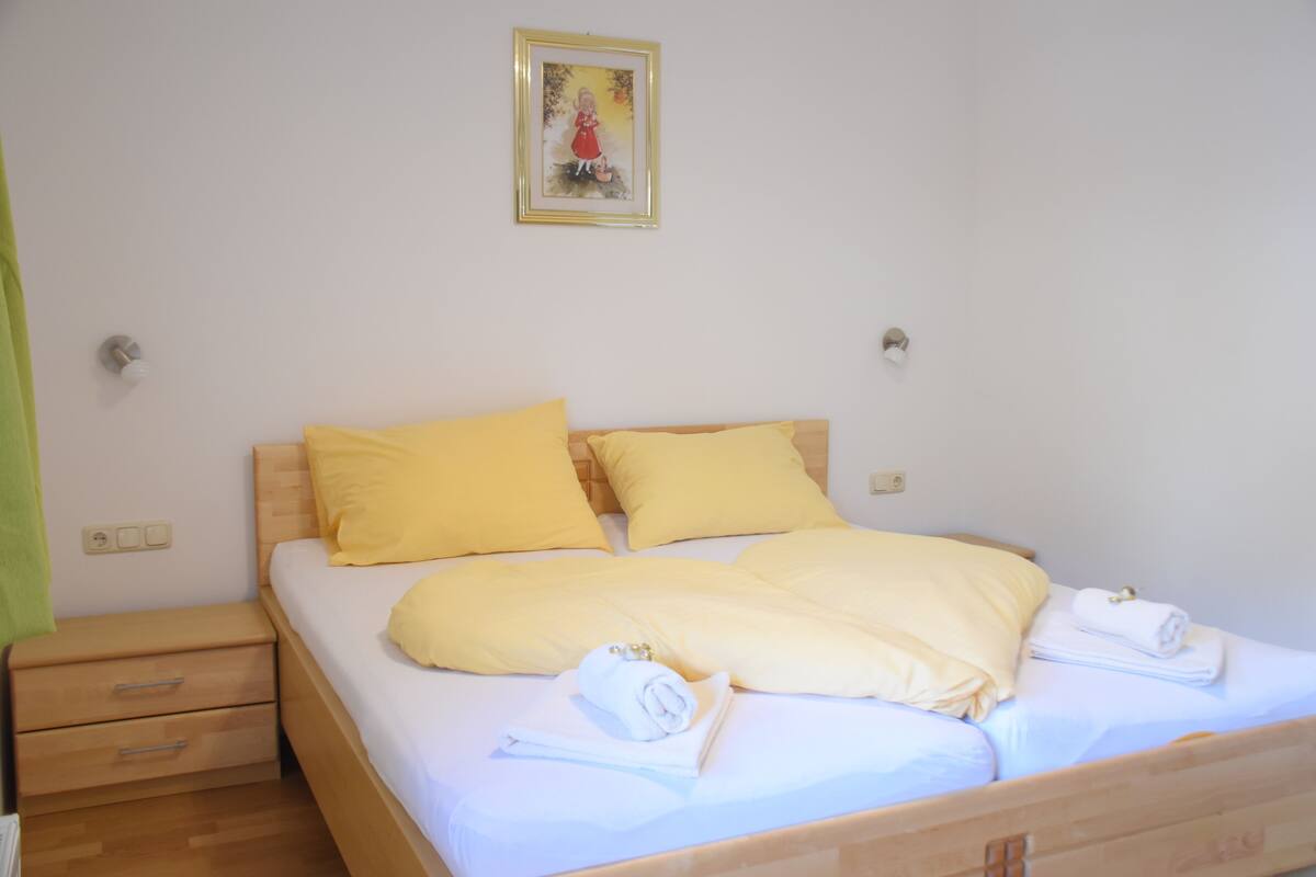 Una cama bien hecha con ropa de cama amarilla y toallas blancas, una mesita de noche de madera con cajones y un cuadro en la pared.