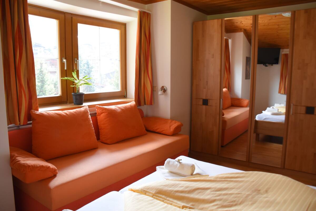 Un dormitorio moderno con sofá cama, un ventanal con cortinas, una planta en maceta y muebles de madera.