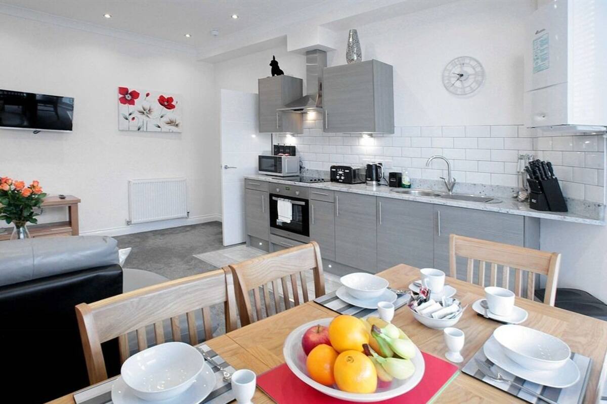 Una cucina moderna con mobili grigi, un forno a microonde e una macchina da caffè. C'è un tavolo da pranzo con un sottopiattino rosso e una ciotola di frutta.