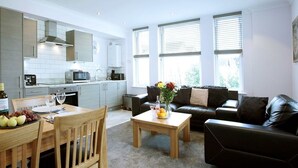 A modern kitchen with white cabinets, a dining area with a wooden table and chairs, and a living room with a black leather sofa and a coffee table. - Austen's Luxury Apartments (Torquay)