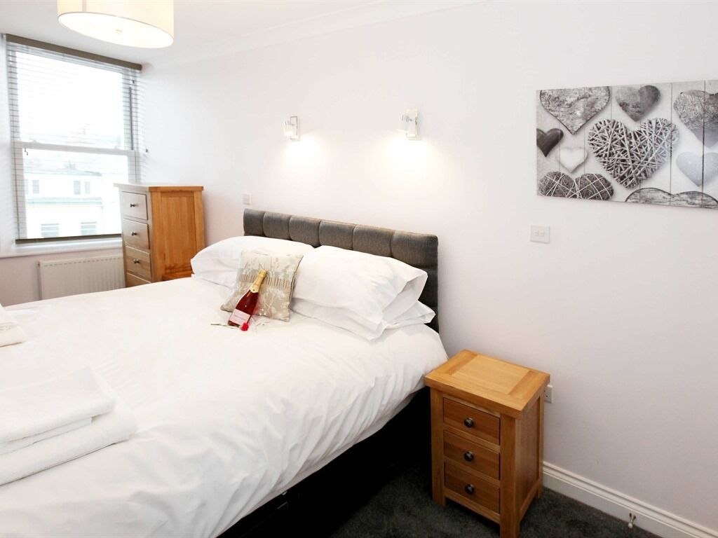 A neatly made bed with white linens, a wooden nightstand with drawers, and a wall-mounted light fixture.